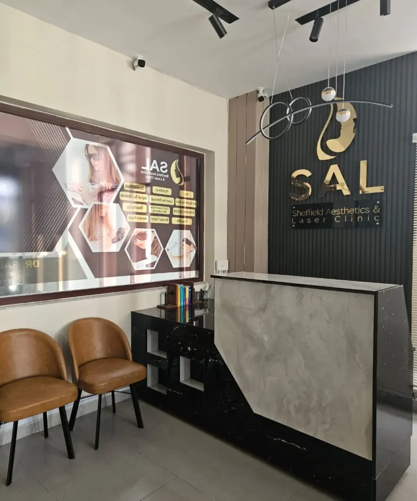 The elegant and modern reception area of SAL Clinic in F7 Markaz, Islamabad, featuring a sleek black and white counter, comfortable leather chairs, and the clinic's gold logo on a dark textured wall. This interior shot showcases the welcoming ambiance of a leading aesthetics and laser clinic in Pakland Tower. Keywords: SAL Clinic, reception, interior, F7 Markaz Islamabad, Pakland Tower, aesthetic clinic, laser clinic, skincare clinic, modern design, luxurious environment.